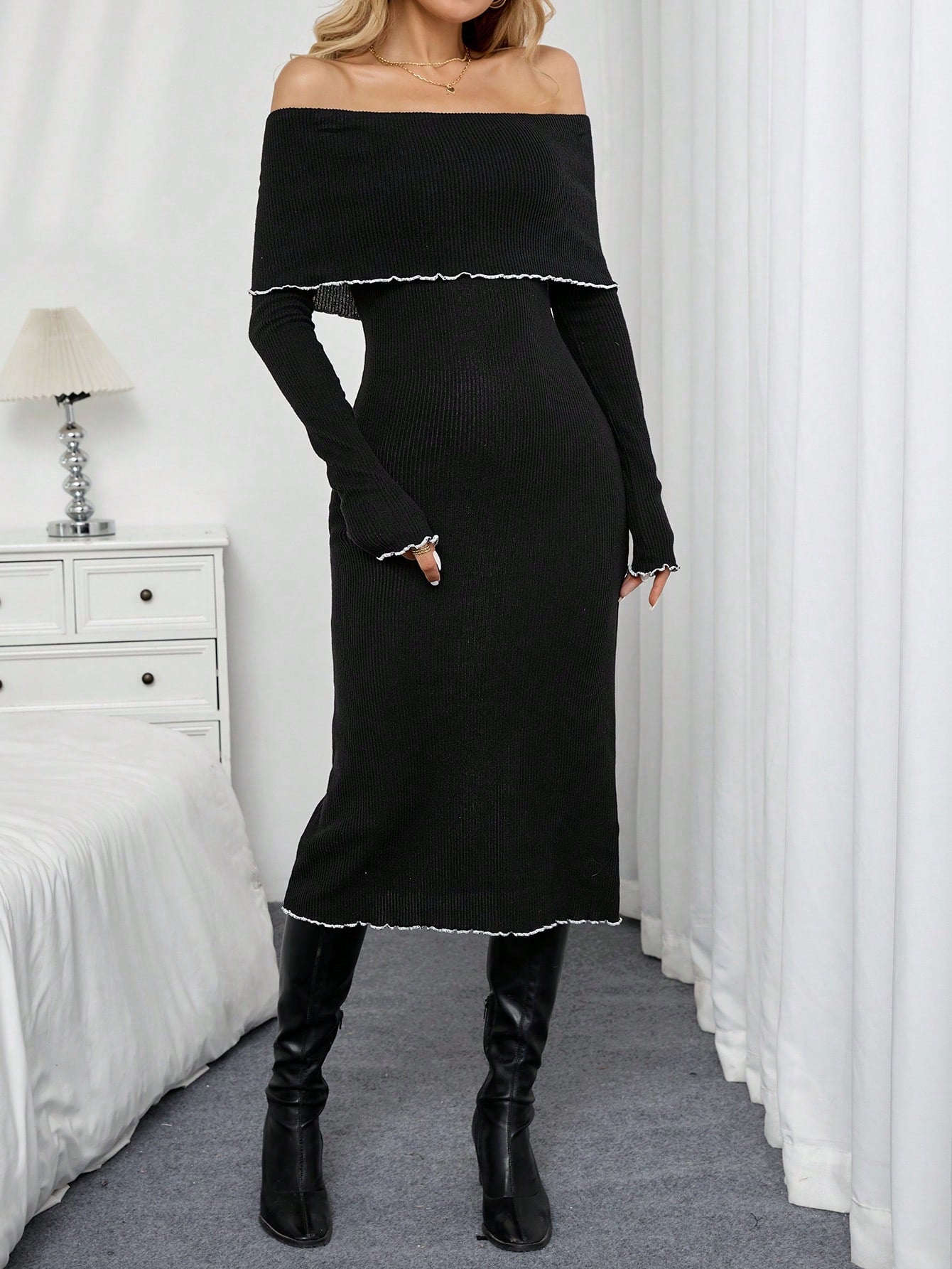 A woman models a black off-the-shoulder long-sleeve midi dress with white scalloped trim, paired with black knee-high boots, standing in a bedroom setting with white curtains and a bed.