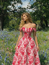 A woman wearing a red and white floral off-the-shoulder ball gown stands in a sunlit outdoor field of blue flowers with trees in the background.