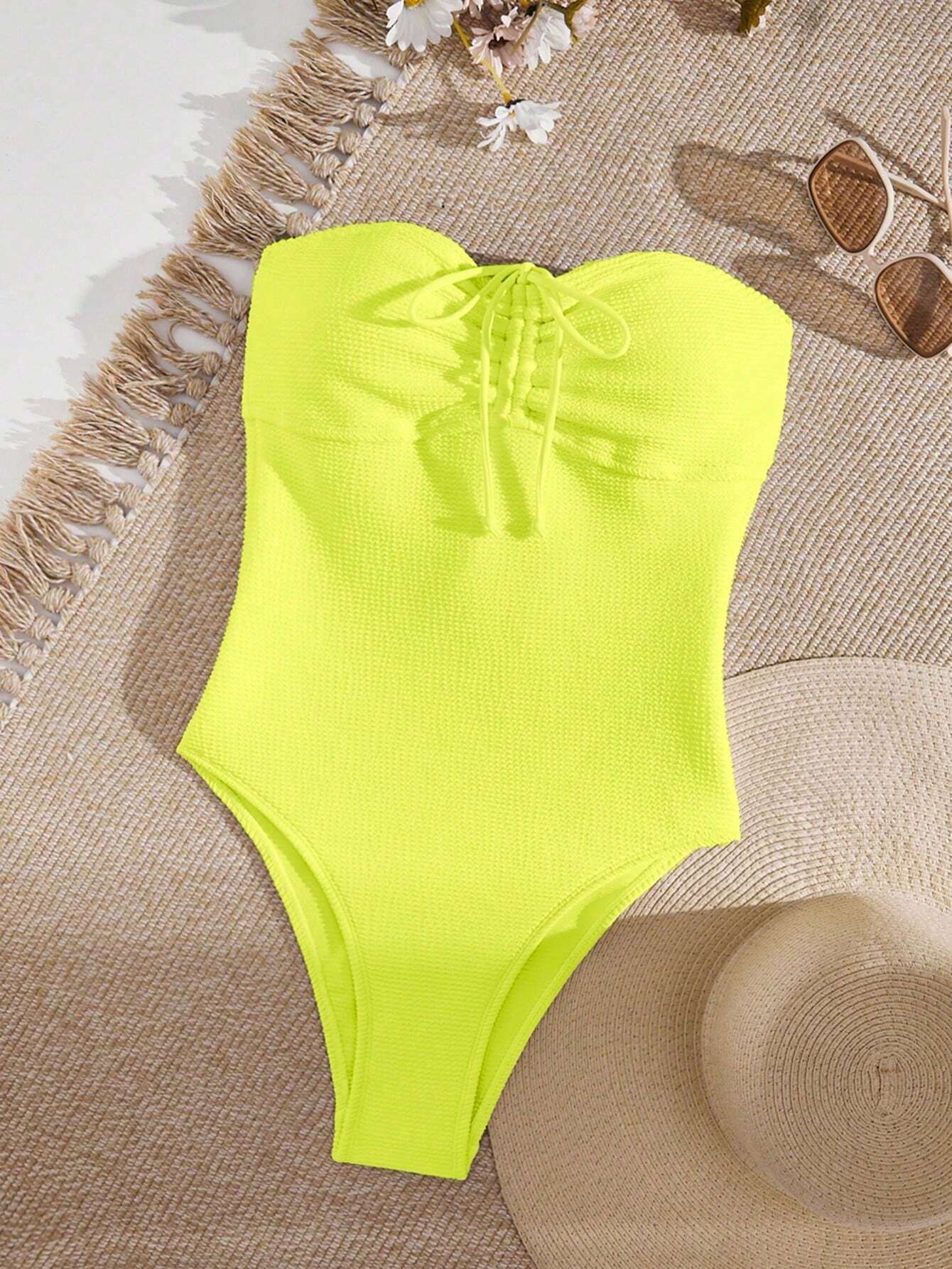 A bright neon yellow one-piece swimsuit laid flat on a beige woven mat with fringe, alongside sunglasses, flowers, and a straw hat.
