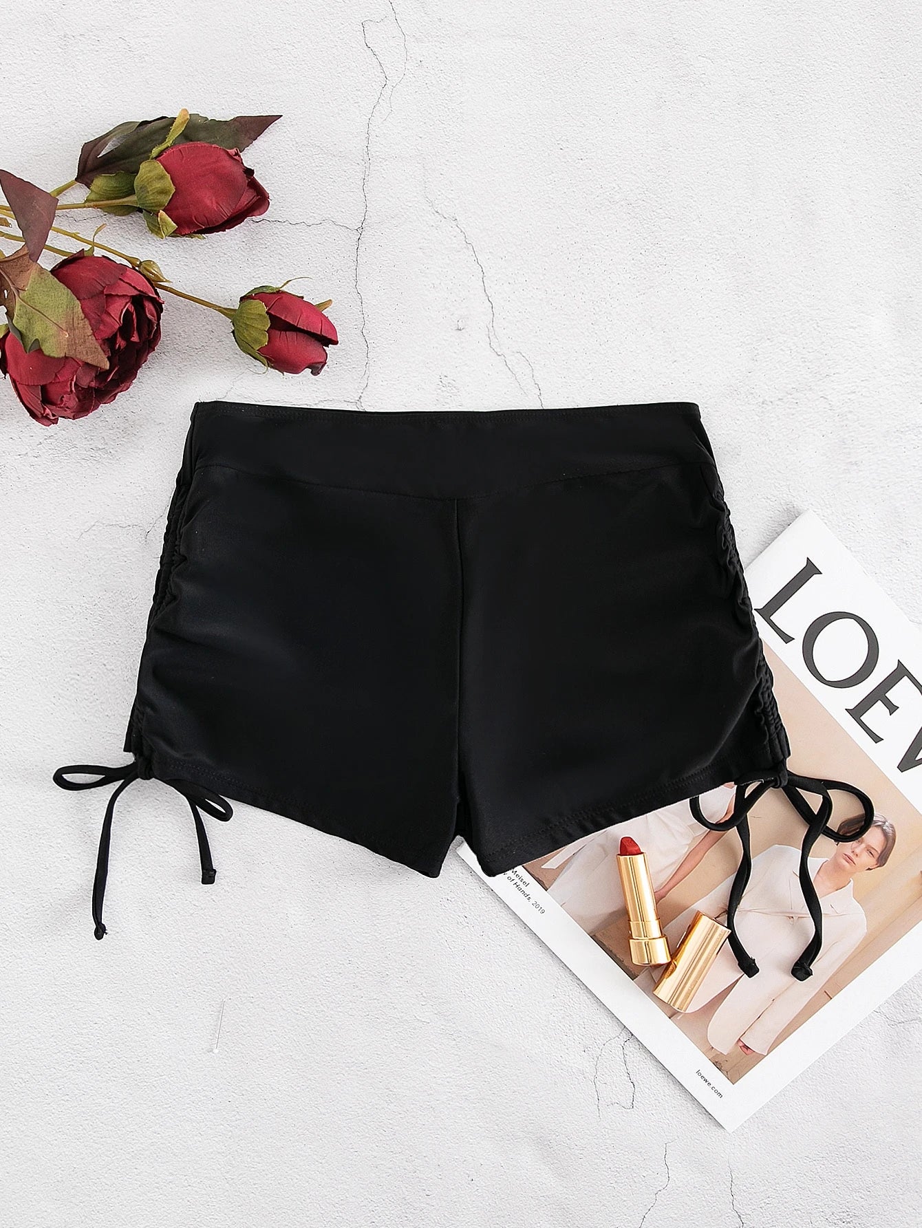 Black high-waisted side-tie shorts laid flat on a light textured surface with red roses and a magazine under part of the shorts.
