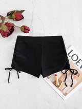 Black high-waisted side-tie shorts laid flat on a light textured surface with red roses and a magazine under part of the shorts.
