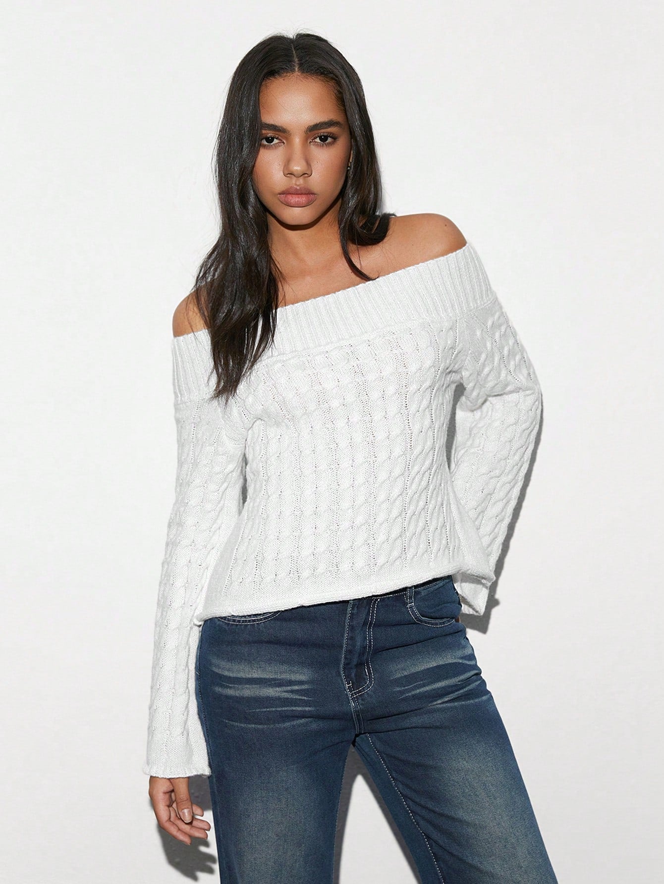 A woman modeling a white off-the-shoulder cable-knit sweater with dark blue jeans, standing against a plain white studio background.