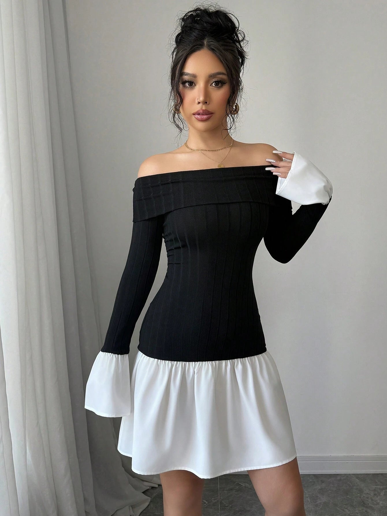 A woman modeling a black off-the-shoulder long-sleeve mini dress with contrasting white bell sleeves and a white skirt, standing indoors against a light gray wall and curtain.