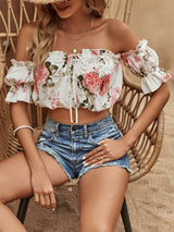 A woman seated on a wicker chair wearing an off-the-shoulder white crop top with a pink floral print and blue denim shorts in a warm neutral indoor setting.