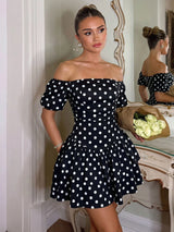 A woman models an off-the-shoulder black dress with white polka dots, standing in a decorated room with a mirror and a bouquet of white roses.