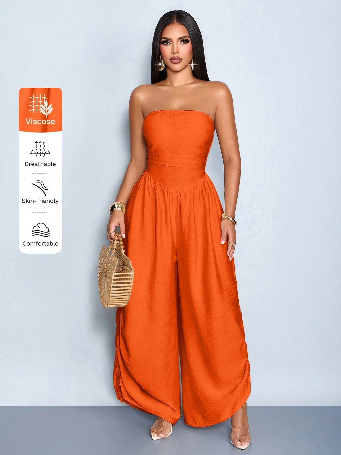 A woman models a bright orange strapless wide-leg jumpsuit against a light blue-gray studio background. She is holding a small woven gold handbag, and a vertical feature callout on the left lists fabric benefits: viscose, breathable, skin-friendly, and comfortable.