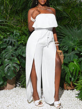 Woman wearing a white off-shoulder dress with a slit, standing in front of green foliage.