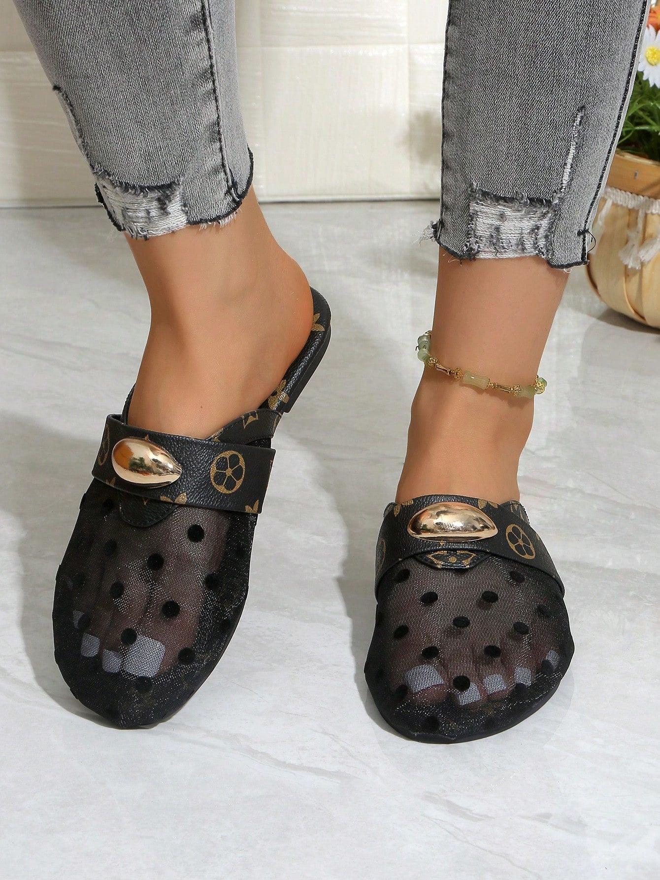 A person wearing black perforated clog-style mule sandals with gold-tone decorative hardware, shown from the calves down against a light indoor floor and wall.