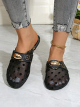 A person wearing black perforated clog-style mule sandals with gold-tone decorative hardware, shown from the calves down against a light indoor floor and wall.