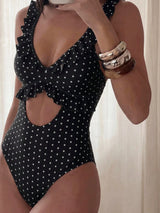 A woman wearing a black one-piece swimsuit with white polka dots, featuring ruffled trim and a side cutout, photographed in a mirror selfie style against a light curtain background.