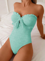 A woman wearing a strapless one-piece swimsuit in mint green with a textured floral pattern, ruffled trim, and a front bow detail, posed on a white bed.