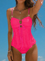 A woman wearing a bright neon pink one-piece swimsuit with thin shoulder straps, a keyhole cutout at the bust, and a ruffled corset-style bodice, photographed against a clear blue sky.
