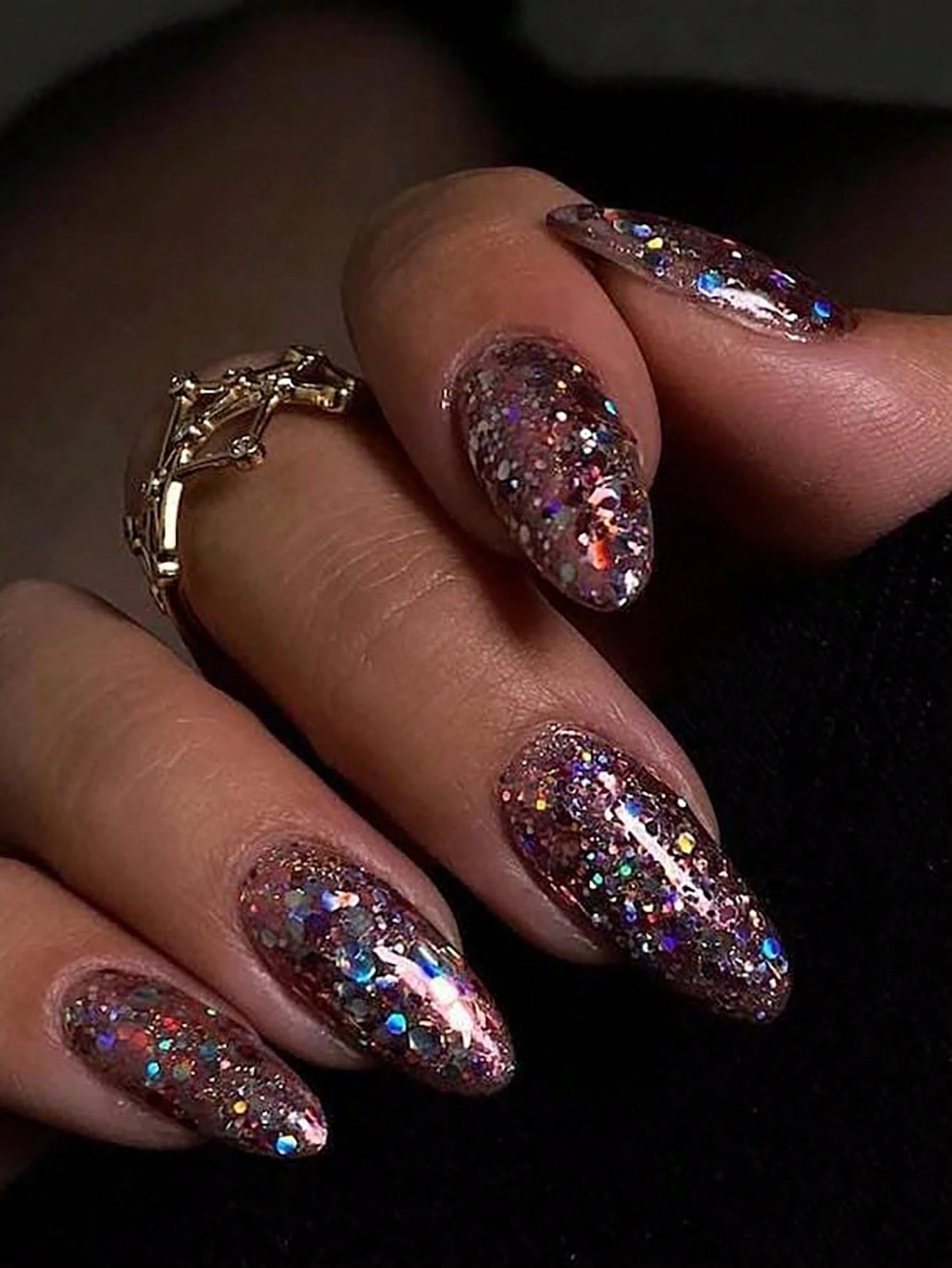 Close-up of a hand with glittery pink nails and a gold ring on a dark background