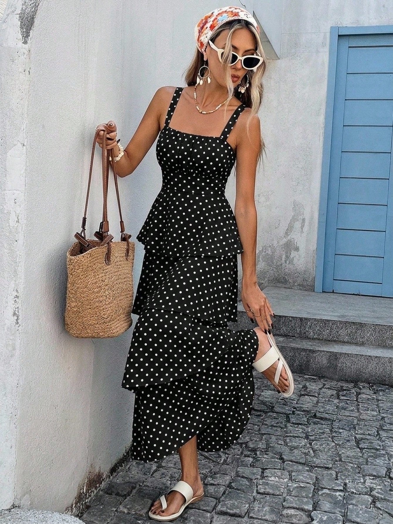 A woman models a black midi dress with white polka dots, styled with a straw tote bag, white slide sandals, a floral headscarf, and sunglasses, standing on a cobblestone street against a white wall.