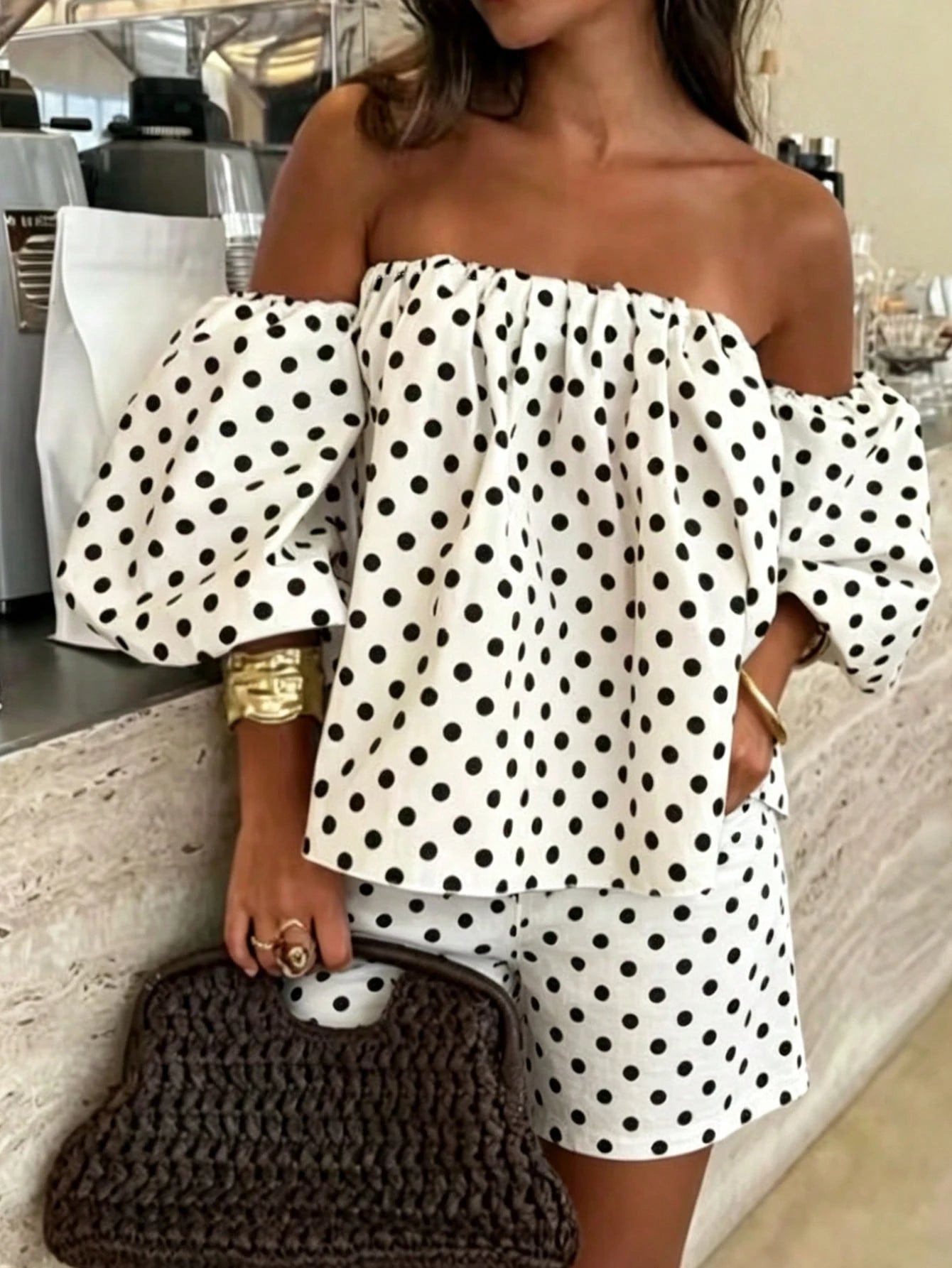 A woman wearing an off-the-shoulder white two-piece set with black polka dots, featuring a loose long-sleeve top and matching shorts, holding a dark brown woven handbag.