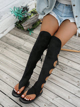 A person seated on a weathered wooden deck wearing black over-the-knee cutout sandals with side laces, light denim shorts, and a light denim jacket.