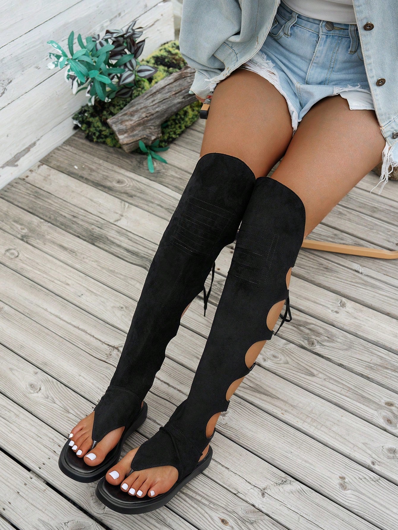 A person seated on a weathered wooden deck wearing black over-the-knee cutout sandals with side laces, light denim shorts, and a light denim jacket.