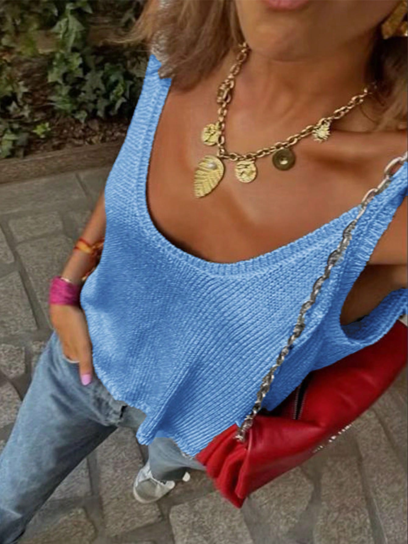 Person wearing a blue top with a gold necklace and red bag on a pavement background