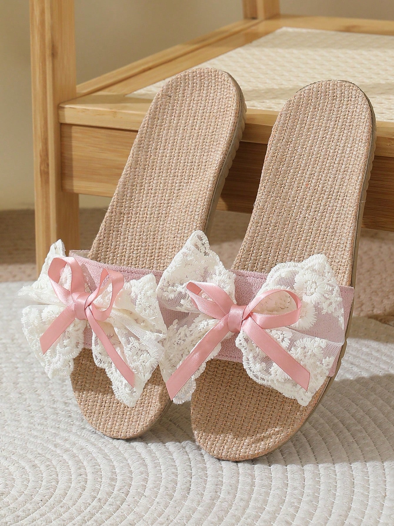 Pair of beige sandals with pink bows and lace details on a wooden bench.