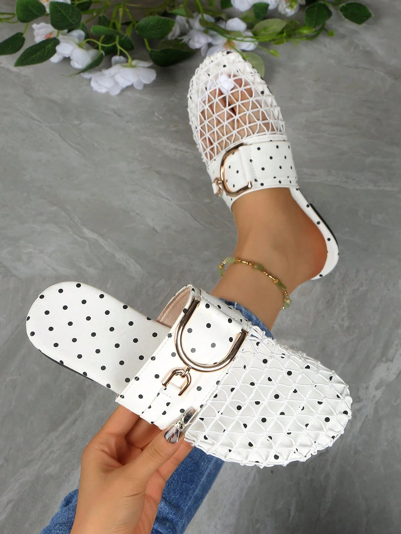 White slide sandals with black polka dots and gold-tone buckle details, shown on a person's foot and held in a hand against a gray stone-textured background with white flowers.