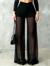 A model wearing high-waisted black sheer mesh wide-leg pants over black sheer tights, paired with a black long-sleeve crop top and black high-heeled sandals, holding a black textured handbag against a gray concrete wall.